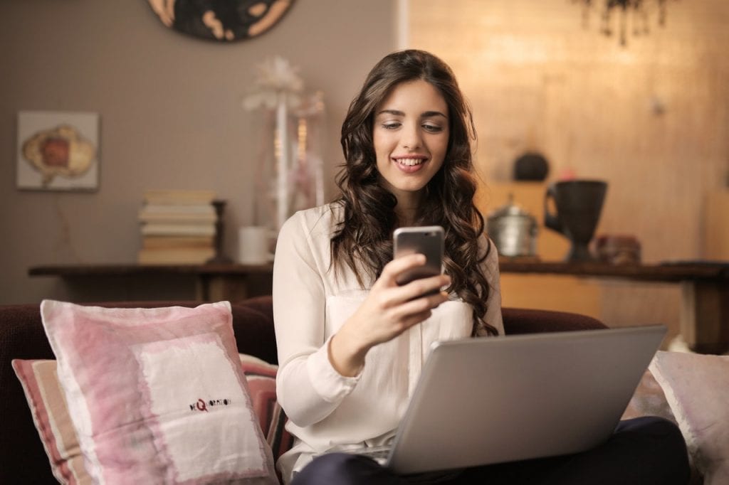 Mulher sentada em um sofá, trabalhando em seu portfolio através de um laptop e um celular em mãos.